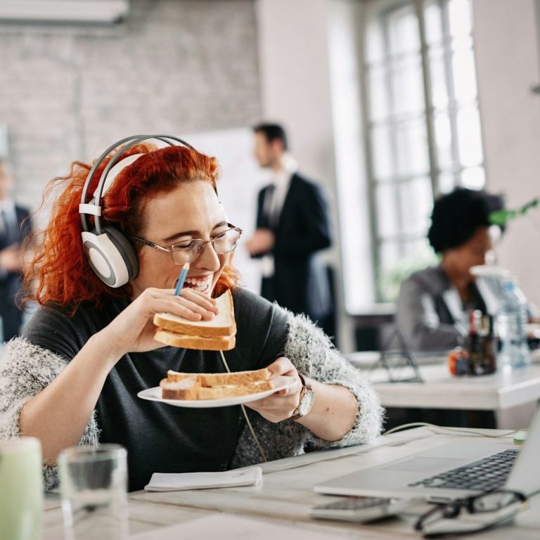 Frau mit roten Haaren und Headset auf dem Kopf, im Büro, isst ein Sandwich